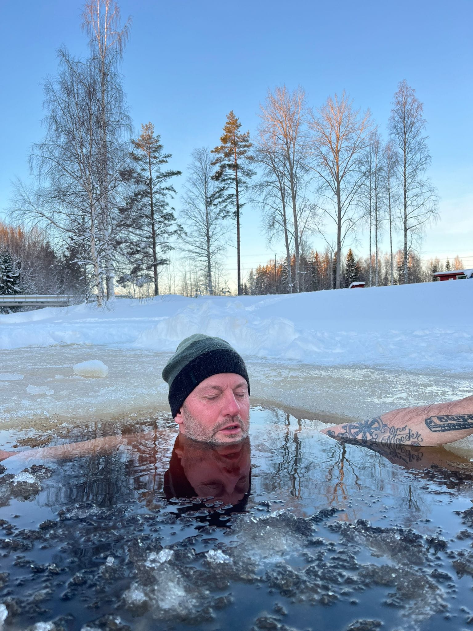 Iemand tot aan de schouders in een gezaagd wak in het ijs, met besneeuwde berken erachter